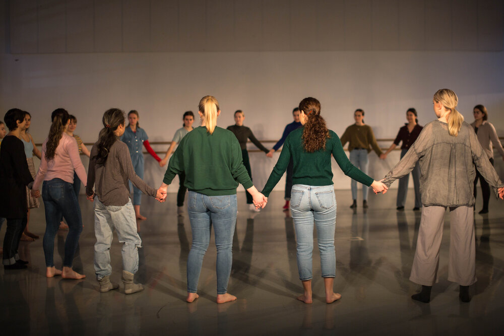 16 dansere står i en stor ring og holder hverandre i hendene